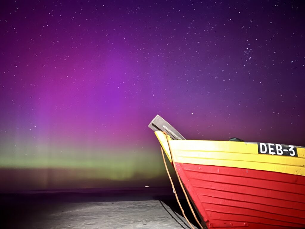 Kolorowa fioletowo-zielona zorza polarna nad plażą w Dębkach z charakterystyczną łodzią rybacką DEB-3 na pierwszym planie.