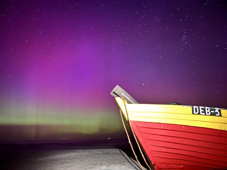 Kolorowa fioletowo-zielona zorza polarna nad plażą w Dębkach z charakterystyczną łodzią rybacką DEB-3 na pierwszym planie.
