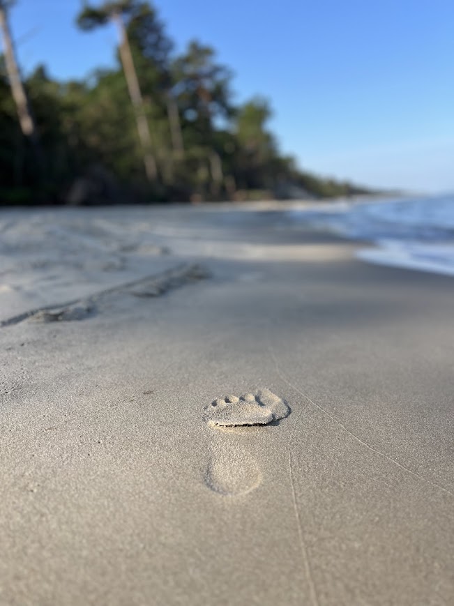 Zbliżenie na odcisk stopy na mokrym, jasnym piasku plaży w Dębkach, w tle rozmyty widok na las i morze