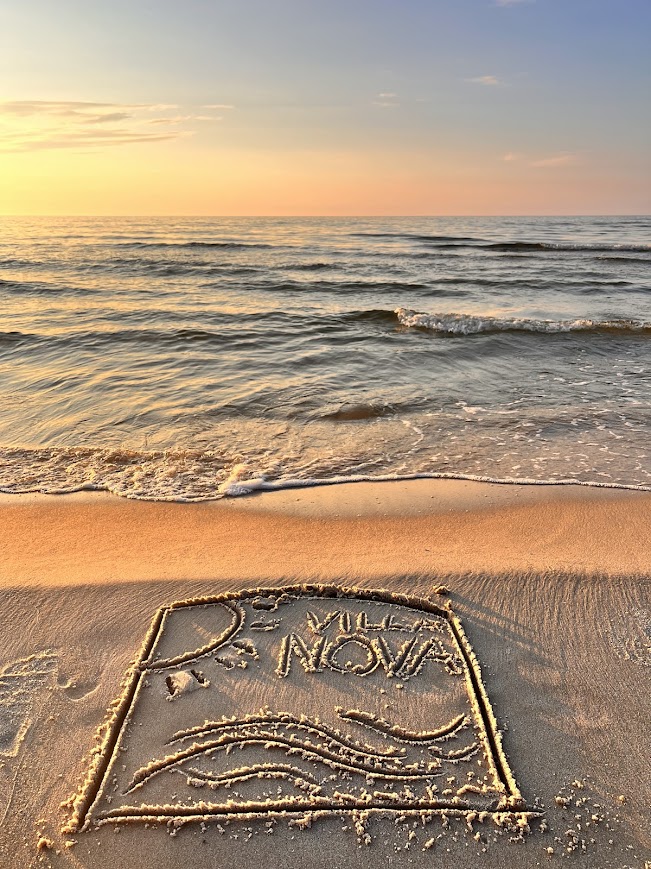 Napis Villa NOVA narysowany na jasnym piasku plaży w Dębkach podczas zachodu słońca nad morzem