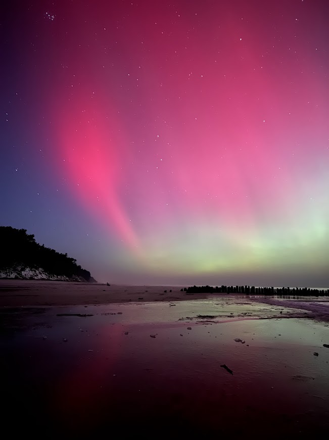 Pionowe ujęcie intensywnej fioletowo-różowej zorzy polarnej nad drewnianymi falochronami i mokrą piaszczystą plażą w Dębkach poza sezonem