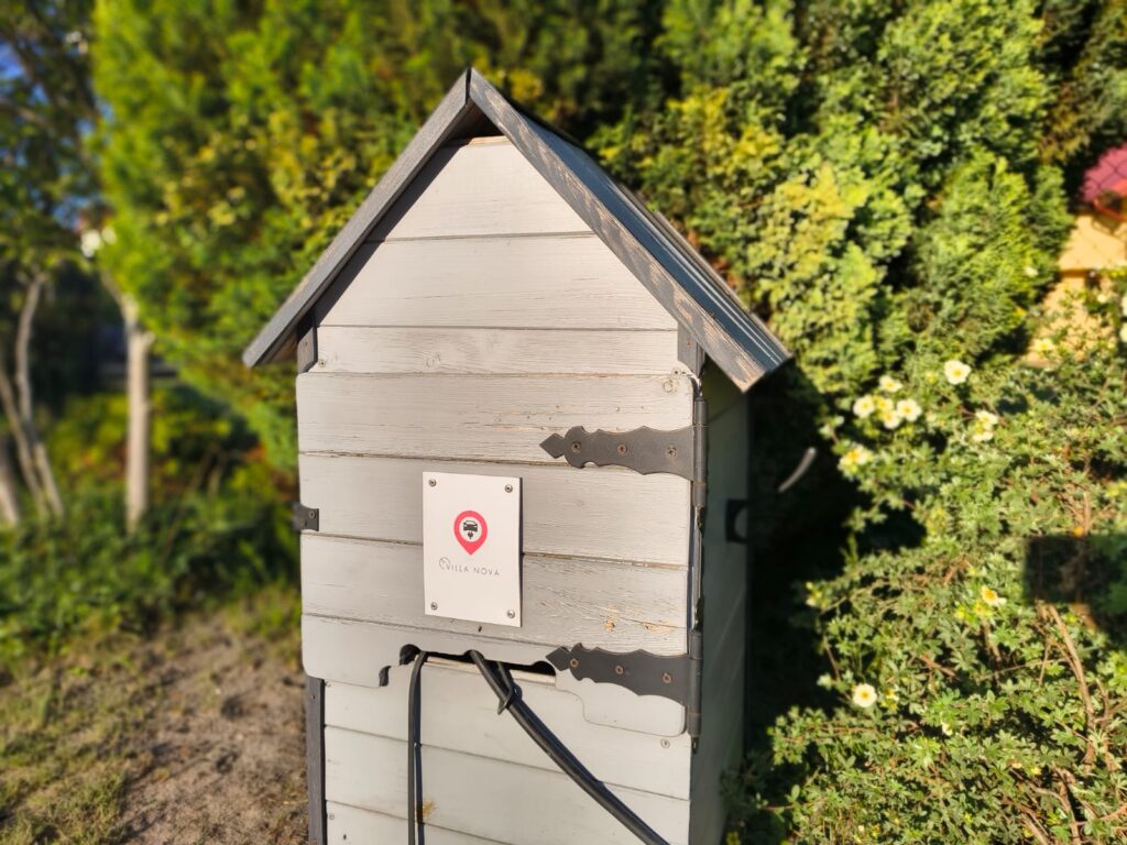 Stacjonarny punkt ładowania samochodów elektrycznych z logo Villa NOVA Dębki w ogrodzie