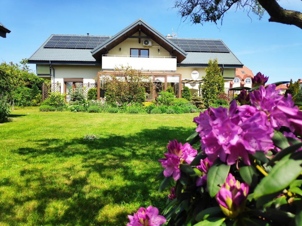 Villa NOVA Dębki – widok na budynek i zielony ogród z kwitnącymi kwiatami